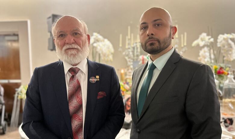 Two men wearing suits look at the camera with solemn expressions.