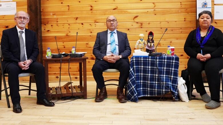 Two people in suits and one person in a sweater and blue scarf sit on chairs. 