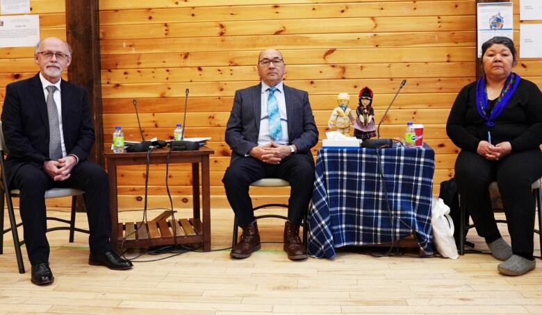 Two people in suits and one person in a sweater and blue scarf sit on chairs. 