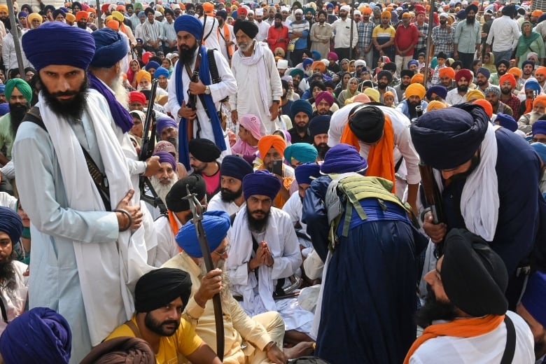 A man wearing a navy blue turban and a long, white scarf over a light-coloured robe, stands on the left of the frame surrounded by a crowd of people sitting on the ground.