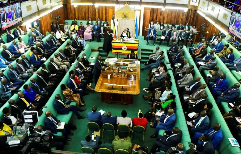 Wide shot of room in legislature with green carpet and breen benches all fully occupied.