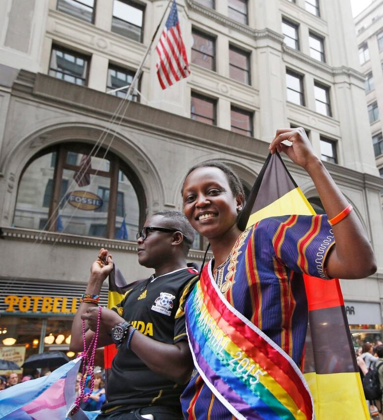 Two individuals standing atop a float in a parade, the woman wearing a colourful dress and sash and smiling 