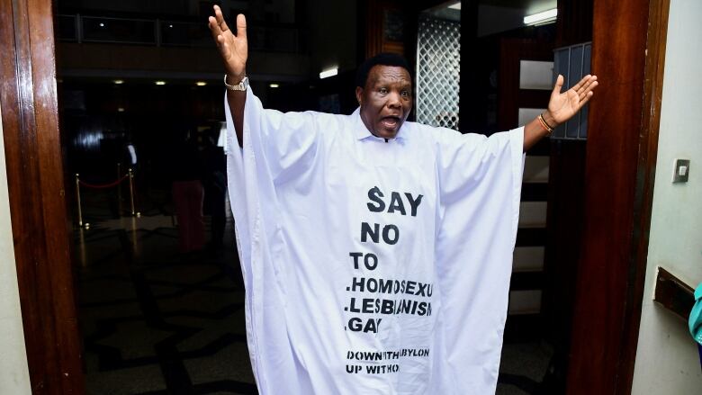 A man raises his hands in the air while wearing a long, white gown with any anti-LGBTQ message on it in black print. 