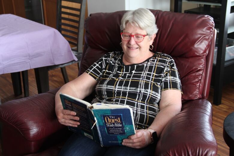 A woman with grey hair and red-framed glasses smiles as she sits in an armchair, reading a slightly tattered book titled 