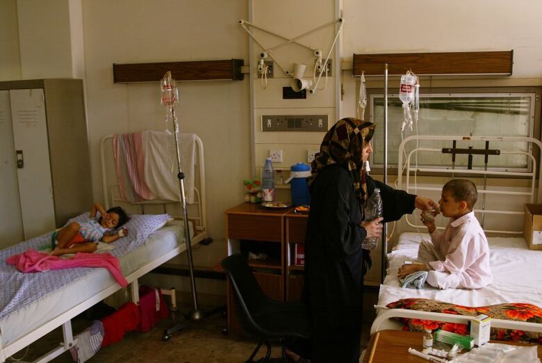 A woman wearing a black robe and a headscarf stands in a hospital room next to a child sitting on a bed.