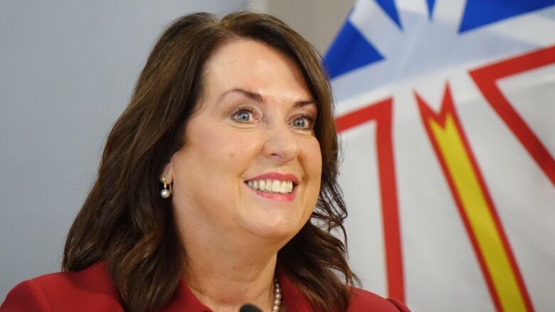 A dark-haired woman wearing a red blazer smiles, standing in front of a flag.