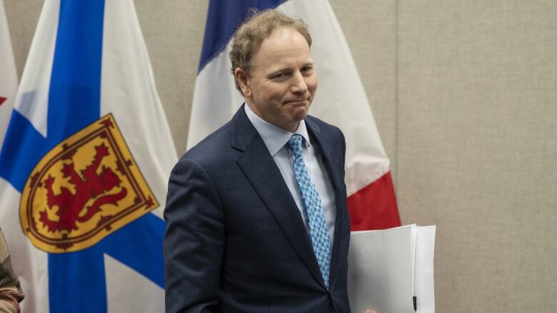 A man in a suit holds a folder of papers, with Nova Scotia flag in the background.