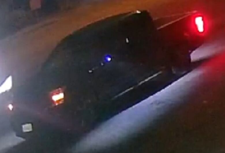 A dark, grainy surveillance photo shows a dark-coloured pickup truck driving with its lights on.