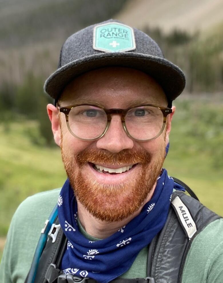 A many in a cap that says Outer Range, with a blue bandana around his neck and wearing glasses, poses for a photo in front of some mountains.