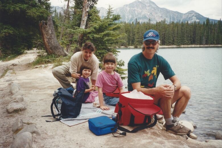 A man and woman sit for a photo with two little girls on the rocks at a lake surrounded by forest. 