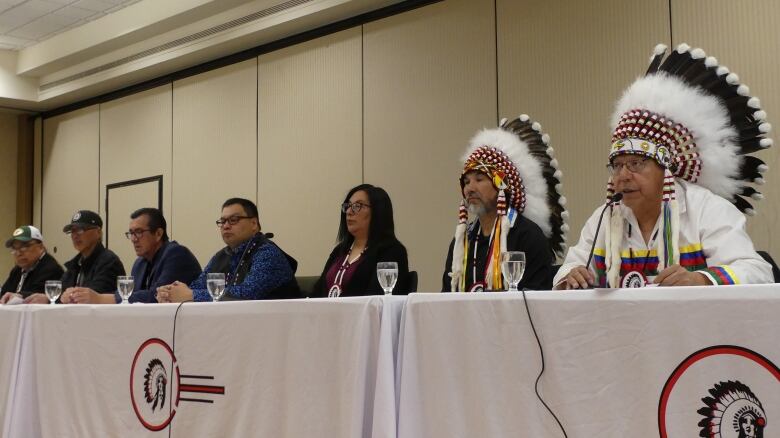 A group of people are lined up on one side of a table. An Indigenous man wearing a headdress speaks into a microphone.