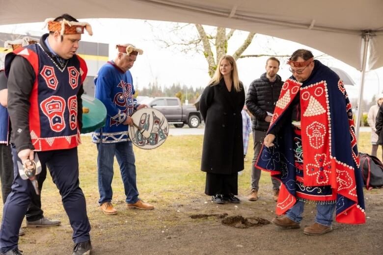 Men in Indigenous clothing and headbands hold drums and are singing a blessing to the land, with an audience overlooking them. 