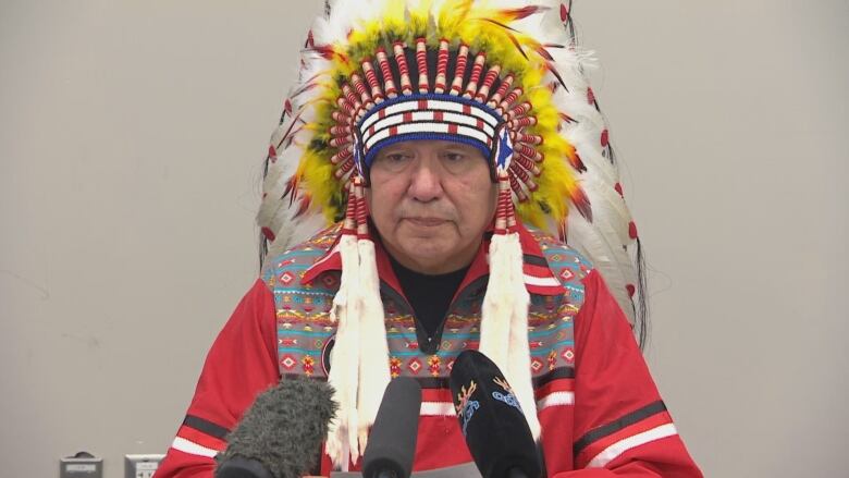 A man in a traditional First Nations headdress made of feathers and beads, and wearing a red shirt with black and white trim, sits at a table and speaks into microphones. 