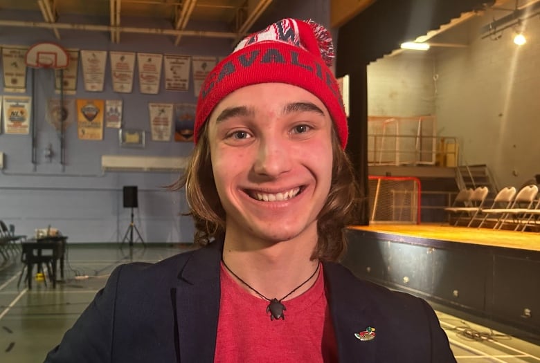 A boy waring a black jacket, red shirt and a winter hat standing in a high school gymnasium. 