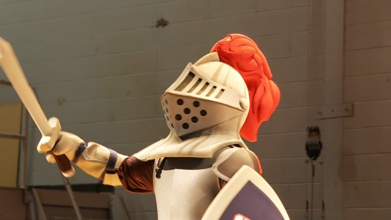 A school mascot dressed in foam armor standing on a stage with a foam sword in its hand. 