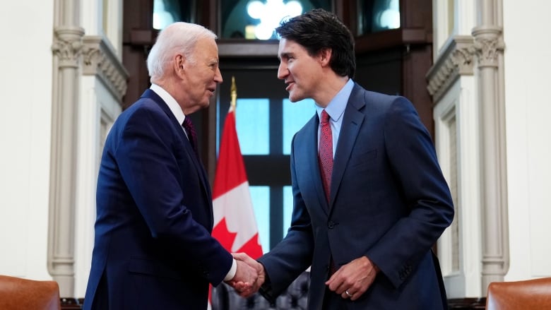 Two men in suits shake hands in an office setting in front of a Canadian flag.