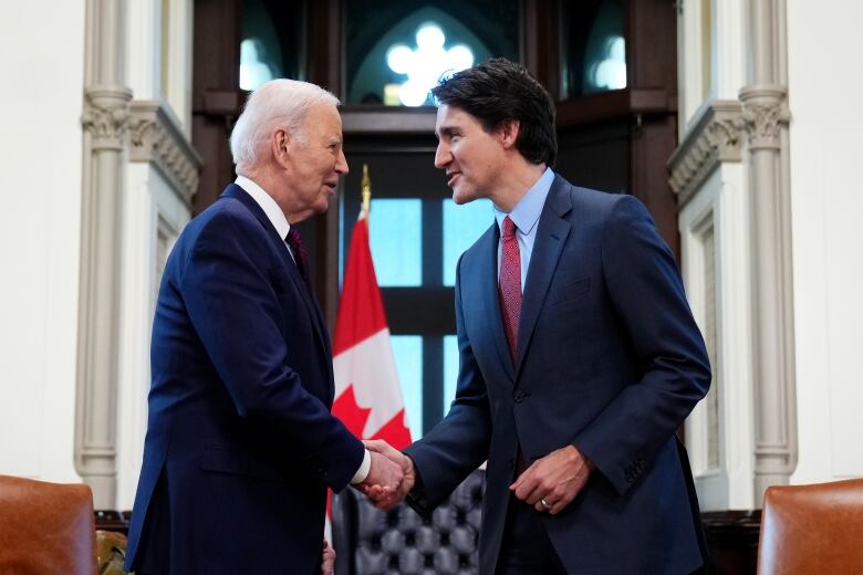 Two men in suits shake hands in an office setting in front of a Canadian flag.