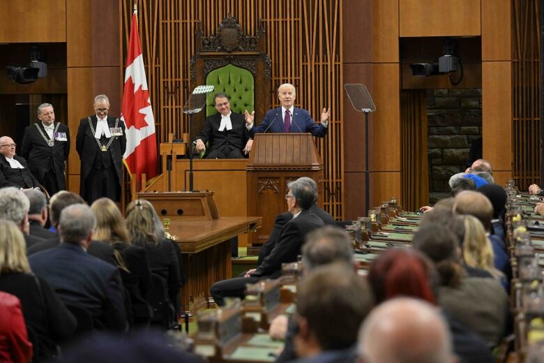 A person gestures while speaking at a lectern as people seated look on.