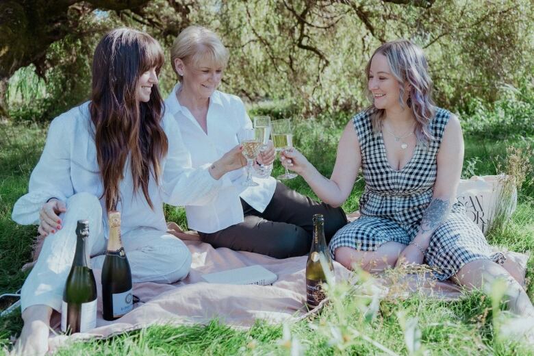 Three women sit on a picnic blanket in a field. They are drinking from wine glasses and smiling.