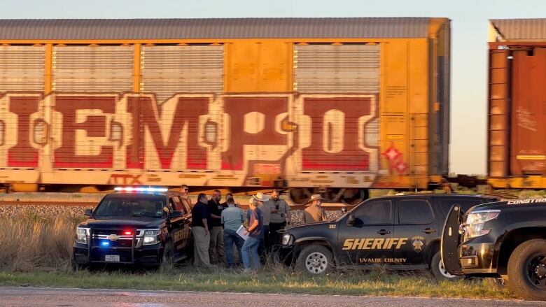 About nine people are seen in the distance standing between two police vehicles and next to a freight train. One of the police vehicles has the words 'Sheriff Uvalde County' on the side. the other has its police lights illuminated.