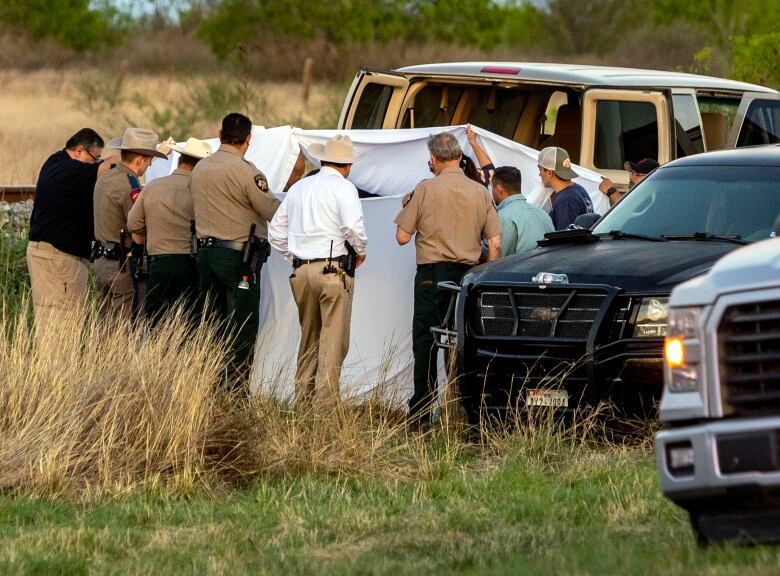 A group of about ten people hold up what appear to be white sheets, likely to obscure the view, as they stand in grassy area. Vehicles are parked near them and a train track can be glimpsed in the background.