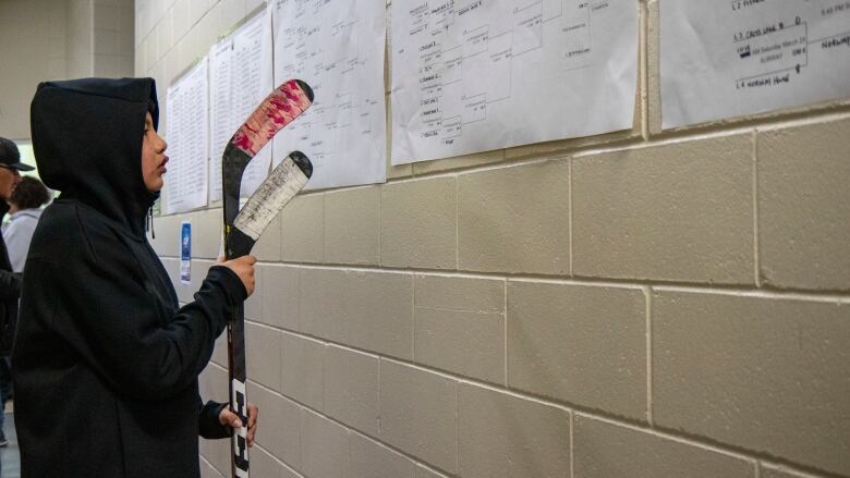 A boy holding two hockey sticks looks at a hockey tournament shedule.