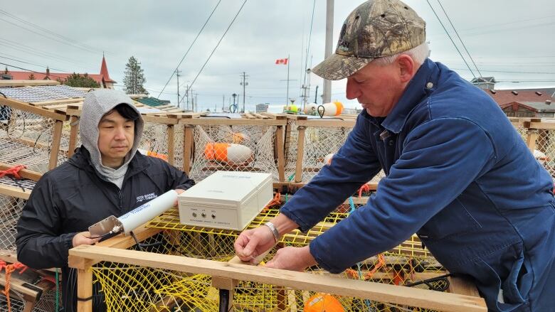 A man wearing a camouflage baseball cap and blue coveralls is attaching a small device to a lobster trap while another man in a hoodie looks on.