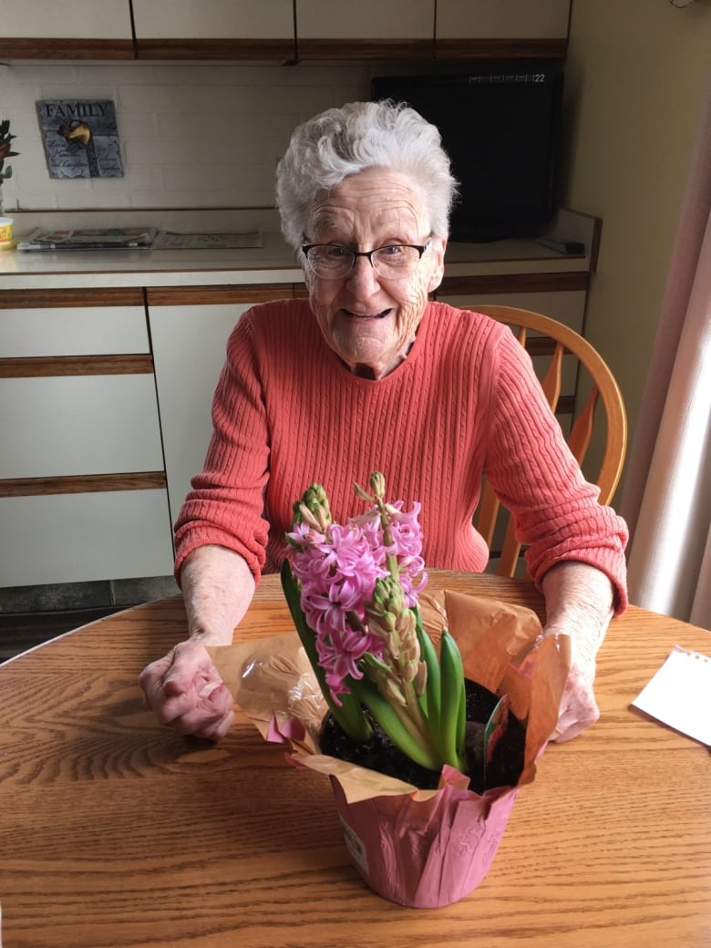 A white-haired woman sits at a table with a pink flower in front of her.