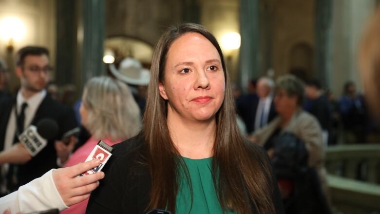 A woman with long hair stands in a legislative building with several other people in the background.