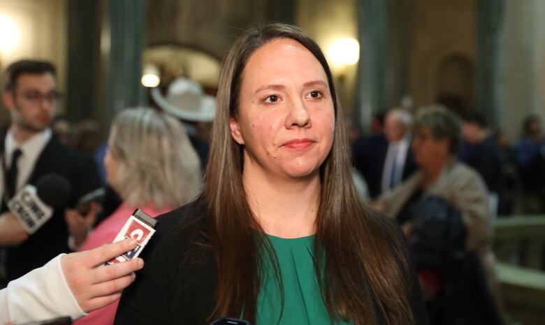 A woman with long hair stands in a legislative building with several other people in the background.