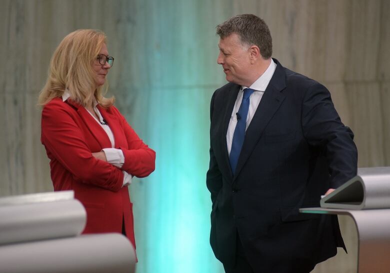 A man and woman in suits stand indoors talking to each other