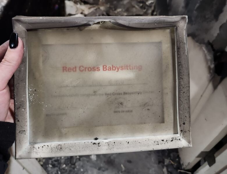 A woman's hand holding a burnt and warped frame that has a babysitting certificate inside of it. 