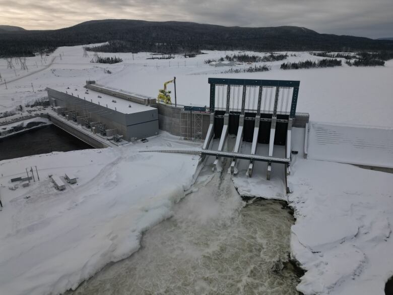 An aerial view of a hydroelectric dam in winter. Water rushes through an open spillway gate.