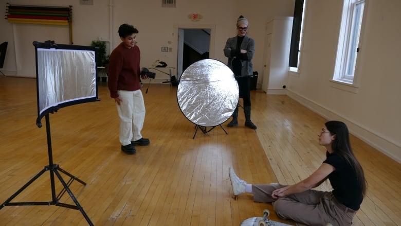 A photographer stands to left in studio with lighting equipment around them. Their assistant stands nearby as a subject with long dark hair sits on the floor with their skateboard