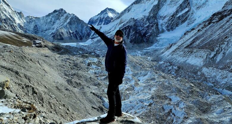 A bearded man pointing up and the left, with snow-covered mountains in the background. 