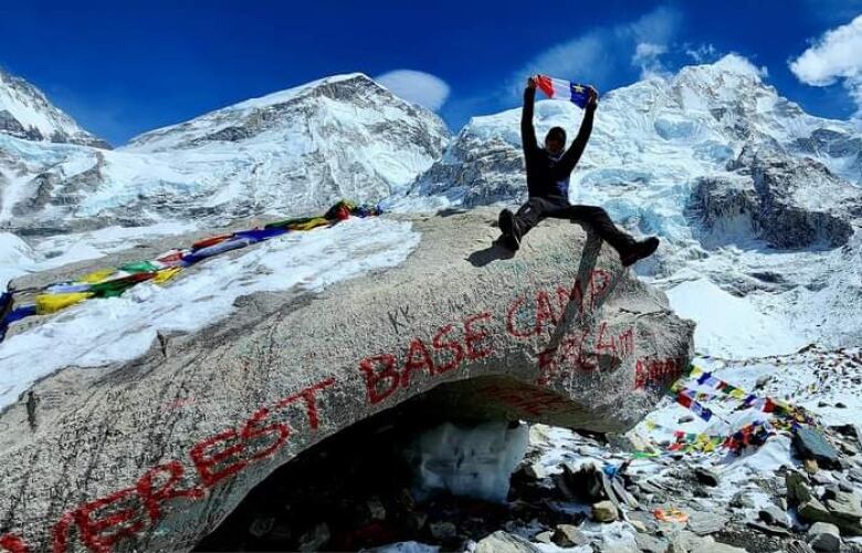 A man sits atop a large rock, lifting a flag over his head, with snow-covered mountains in the background. 