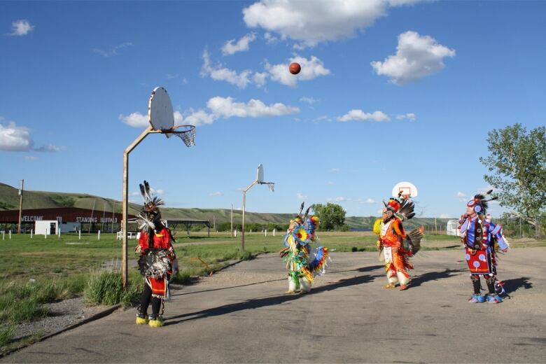 Four people are seen playing basketball on an outdoor court. They are each wearing colourful, traditional Indigenous outfits. 