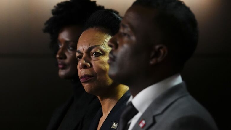 Three black people at a news conference stand in a row and look forward.