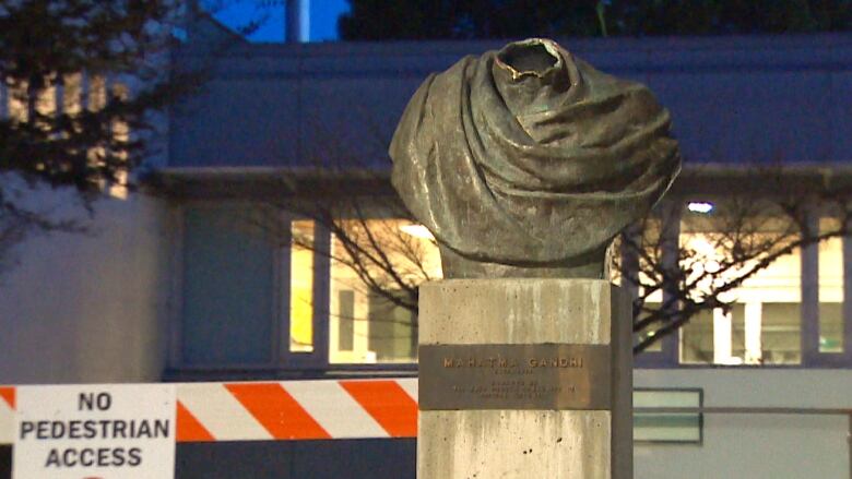 A nighttime, outdoors image shows a concrete pedestal with a bronze nameplate holding the decapitated bust of a statue, with a hollow hole in the neck where the head should be. The shoulders are sculpted to look like they're draped in loose fabric. A building, trees and a traffic barrier can be seen in the background.