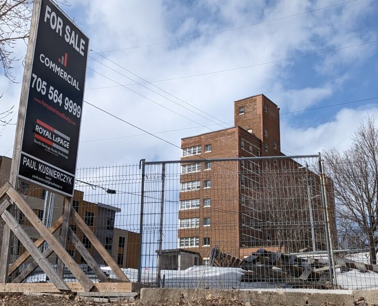 A for sale sign stands outside a brown six-story building with a metal fence around it.