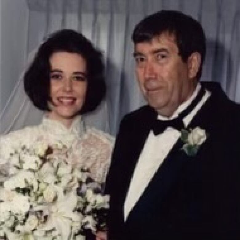 A young woman, in a wedding gown, with a veil and a bouquet, smiles. An older man in a suit, with a boutonniere attached, is standing next to her.