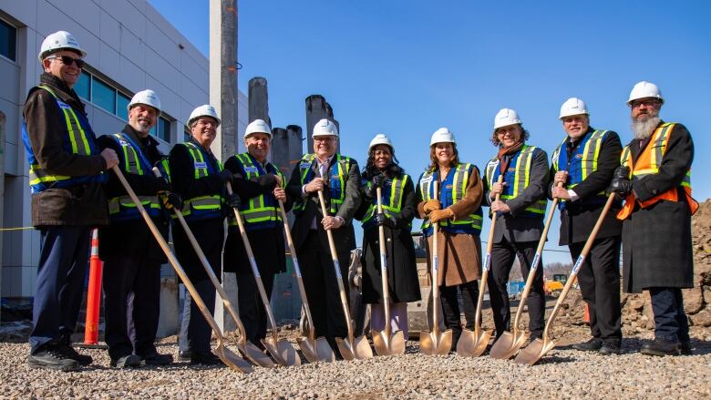 A group of 10 people wearing hard hats and holding shovels stand outside, smiling.