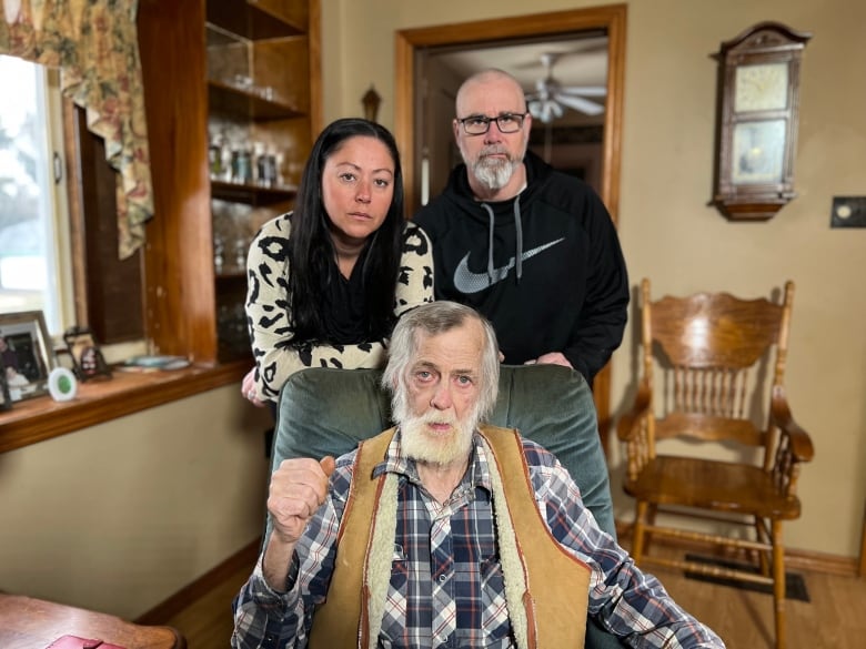 An elderly man sits in a chair while a couple stands behind him. 