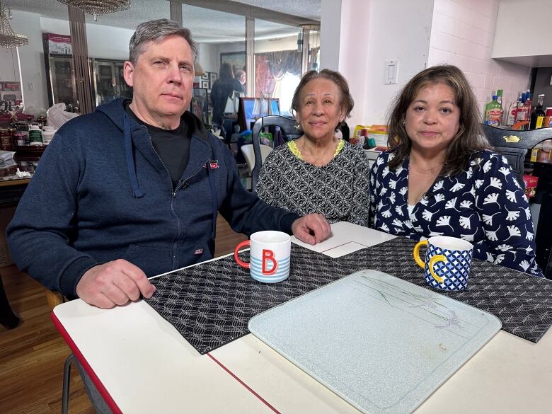 A mother, daughter, and her daughters husband sit at a kitchen table, looking into the camera. 