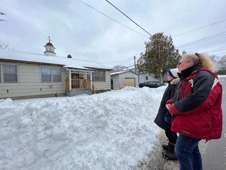 Two women stand on a snowy road looking at a house. 