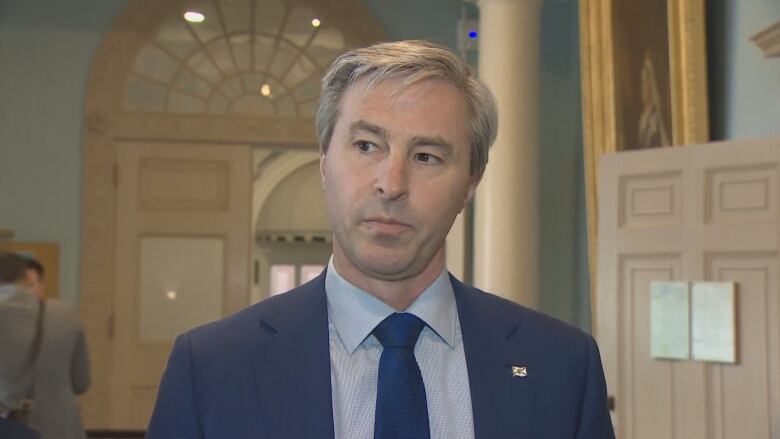 A man with short grey hair wears a blue blazer and blue tie with a light blue shirt.