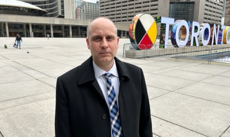 Man in a black coat stands in front of City Hall and the City of Toronto sign.