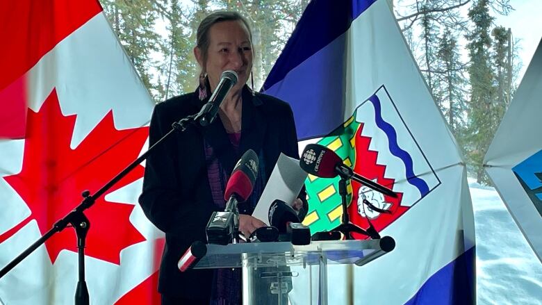 A woman stands smiling at a podium with microphones from the press in front of her and a Canadian flag and Northwest Territories flag behind her.