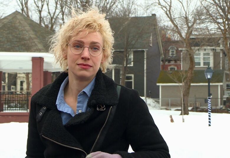 A person with curly blonde hair is outside. Snow and houses can be seen behind them.