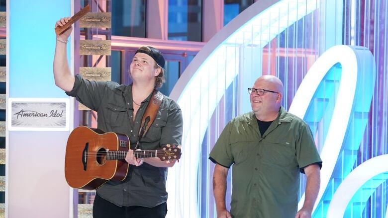 A young man with a guitar stands next to a man with glasses.